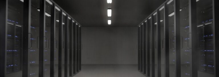 Ciemne pomieszczenie serwerownii. Szafy typu RACK rozmieszczone po lewej i prawej stronie. Datacenter w ciemniej kolorystyce. W tym przypadku serwery i klimatyzatory powodują generację mocy biernej.